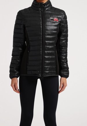 Femme portant une veste matelassée noire ajustée avec des panneaux latéraux, une fermeture éclair frontale, des poches zippées et un logo montagne rouge et noir sur la poitrine.