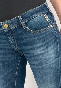 Jeans de mezclilla azul oscuro con un ligero desgaste, que cuentan con un botón dorado, cinco bolsillos y una etiqueta de logo en la parte frontal. Diseño ajustado.