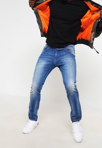 Světle modré džíny z denimu s úzkým střihem a efektem vyblednutí, doplněné černým tričkem a maskáčovou bomber bundou s oranžovým podšívkou.