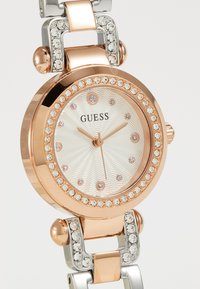 Montre métallisée rose doré et argentée avec un cadran blanc texturé, des accents en cristal et un boîtier rond avec une lunette décorative.