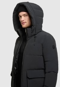Schwarze Steppjacke mit großer Kapuze, Frontreißverschluss, elastischen Kordelzügen und einer Seitentasche mit Klappe. Glatte, matte Textur.