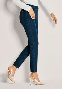 Frau trägt dunkelblaue Skinny-Jeans, ein weißes, strukturiertes Langarmoberteil und weiße Slingback-Pumps mit Mesh- und Perlendetails.