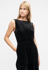 Νεαρή γυναίκα με μακριά ξανθά μαλλιά, φορώντας αμάνικο μαύρο φόρεμα βελούδο με τα χέρια στις τσέπες, κοιτάζει προς τα αριστερά της, σε λευκό φόντο.