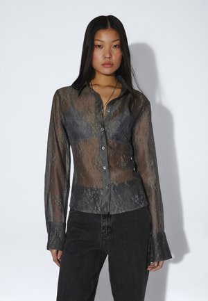 Ung kvinde iført en gennemsigtig grå blomsterbluse over en sort bralette og sorte højttaljede jeans, poserer mod en ensfarvet hvid baggrund.