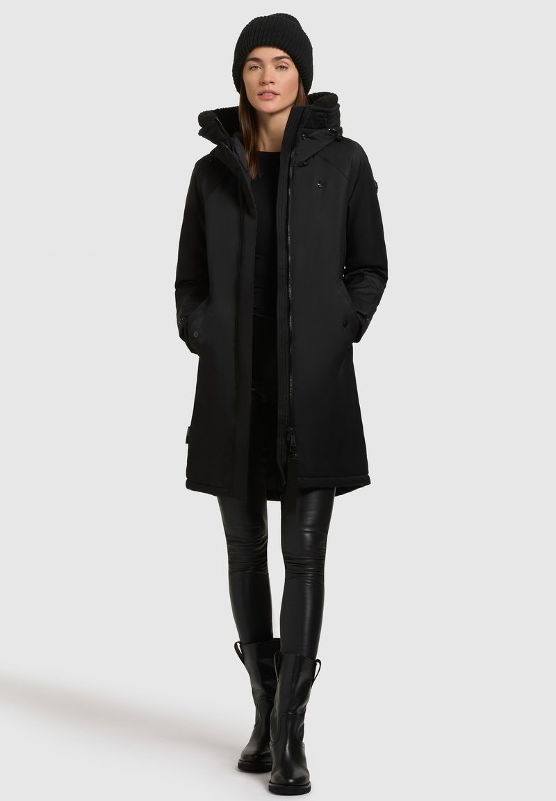 Zwarte waterdichte parka met een capuchon, ritssluiting en zijzakken. Gedragen over zwarte leggings en enkellaarsjes, met een strakke, minimalistische uitstraling.