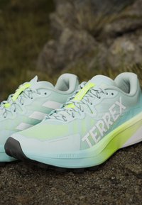 Paire de chaussures de course Adidas Terrex bleu clair et vert néon sur du gravier avec un logo Terrex bien visible sur le côté et des semelles texturées.