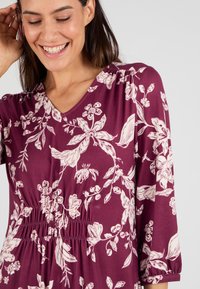 Femme souriante avec de longs cheveux foncés touche sa tête, portant une robe à manches longues bordeaux avec des motifs floraux blancs et une taille froncée.
