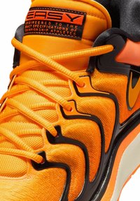 Orange athletic shoe featuring textured mesh material, black accents, distinctive lacing system, and a reinforced sole design.