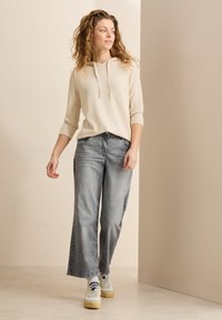 Sweatshirt beige à capuche avec des manches trois-quarts, associé à un jean bleu clair à jambes larges et des baskets blanches à semelles jaunes.