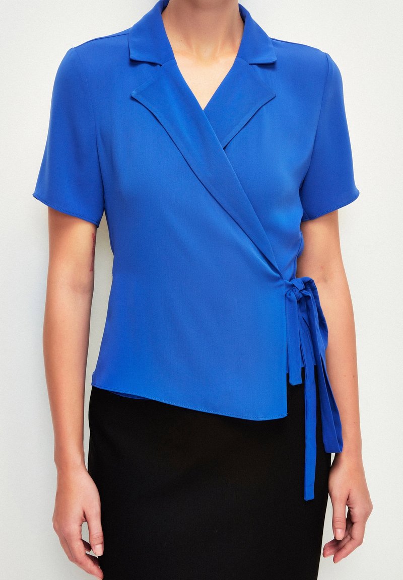 Femme portant un blouse bleue à manches courtes avec col et nœud sur le côté, associée à une jupe noire, torse et bras visibles.