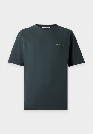 Homme debout les mains dans les poches portant un t-shirt à manches courtes vert foncé, un pantalon noir et des chaussures noires sur fond blanc.