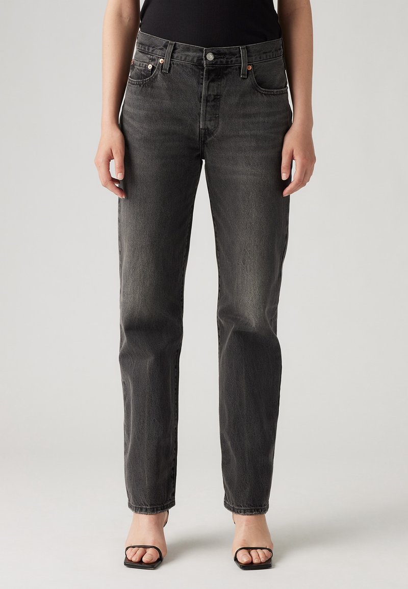 Vaqueros de denim negro con corte recto, cintura de altura media, herrajes de cobre y sutiles desgastes. Costuras blancas acentúan los bolsillos.