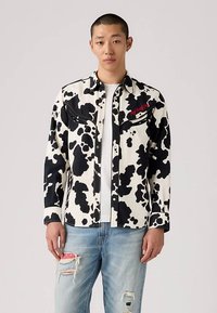 Νέος άνδρας που φοράει ένα πουκάμισο με μαυρόασπρη εκτύπωση γελάδας πάνω από ένα άσπρο μπλουζάκι και ελαφρώς φθαρμένα ανοιχτόχρωμα τζιν με ροζ μπα patch.