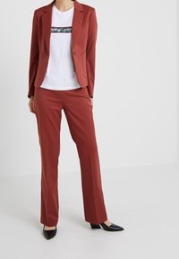 Blazer couleur rouille et pantalon assorti en tissu lisse ; associé à un t-shirt blanc avec un motif graphique. Des escarpins noirs à bout pointu complètent le look.