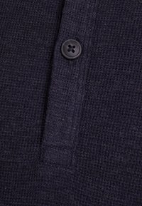 Navy waffle-knit fabric polo with a textured surface and a single black button on the placket, showing detail in the weave and stitching.