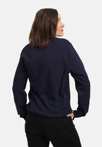 Donna con capelli castani fino alle spalle, che indossa una maglietta a maniche lunghe blu navy e pantaloni neri, in piedi con le mani sui fianchi, guardando di lato.