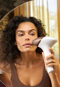 Handheld gezichtsapparaat met een wit lichaam en een ronde bronzen kop. De gebruiker houdt het apparaat dichtbij hun gezicht in een goed verlichte omgeving.