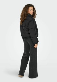 Kvinde med krøllet hår iført en sort dunjakke og sorte bredbenede jeans, stående og kigger over sin skulder mod en ensfarvet baggrund.