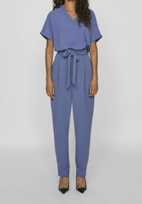 Top blu con scollo a V e maniche corte abbinato a pantaloni a vita alta e su misura, cincti in vita con una cintura; scarpe con tacco nere.