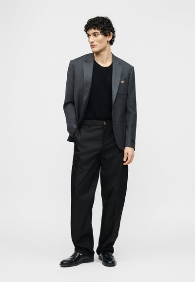 Homme aux cheveux bouclés portant un blazer gris sur une chemise noire, un pantalon cargo noir et des chaussures noires, debout avec une main dans la poche.