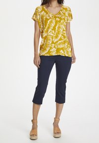 Keltainen lyhythihainen paita, jossa on valkoinen kukkakuosi ja V-pääkappale, yhdistettynä laivastonsinisiin capri-housuihin. Korkeat vaaleat alustakengät, joissa on nilkkahihnat.