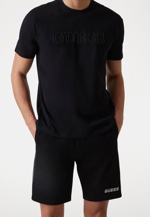 T-shirt noir en coton avec le logo "GUESS" en relief, associé à un short noir arborant un logo imprimé. Les deux pièces ont une texture lisse.
