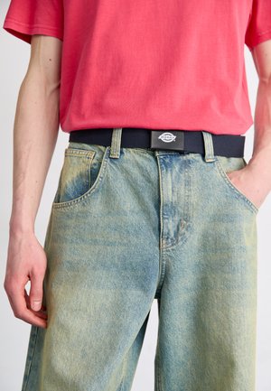 Hellblaue Denim-Jeans mit einer verwitterten Textur, kombiniert mit einem roten Baumwoll-T-Shirt. Ein schwarzer Gürtel mit einem silbernen Logo betont die Taille.