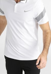 Bílá polokošile s krátkým rukávem a šedými akcenty na rukávech a černým límcem; na levé části hrudi je šedé logo Nike.