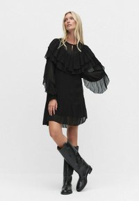 Kobieta ubrana w czarną sukienkę z warstwami falbanek, z przezroczystymi bufiastymi rękawami i wysokimi na kolana czarnymi skórzanymi botkami, pozująca na tle jednolitego tła.