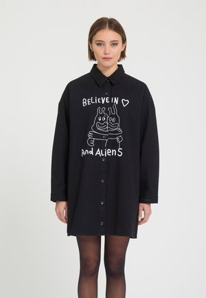 Jeune femme portant une robe-chemise noire boutonnée avec une illustration blanche d’extraterrestre et le texte "Believe in and Aliens" sur le devant.