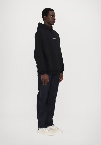 Haut à capuche noir avec poche avant, arborant un texte blanc minimaliste. Associé à un jean bleu foncé et des baskets blanches, se tenant devant un fond uni.