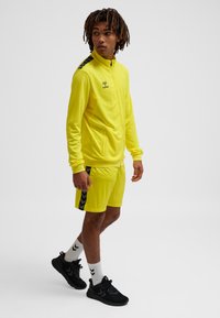 Lyse gul atletisk træningsdragt med sorte accenter; lynlåstrøje, korte ærmer og matchende shorts med sidelængder. Sorte sneakers.