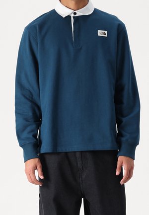 Mann trägt langärmliges marineblaues Rugbyshirt mit weißem Kragen und The North Face-Logo auf der linken Brust, kombiniert mit schwarzen Hosen.