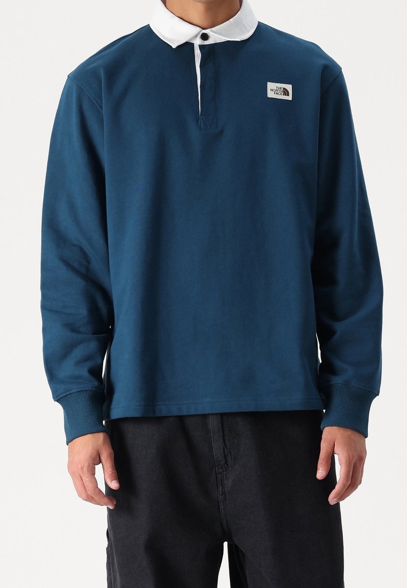 Homme portant un maillot de rugby bleu marine à manches longues avec col blanc et logo The North Face sur le côté gauche de la poitrine, associé à un pantalon noir.