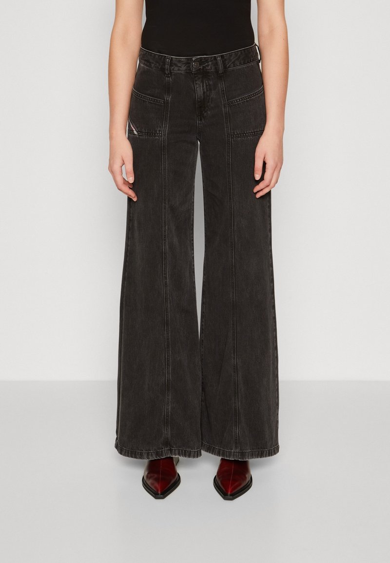 Personne portant un jean noir taille haute à jambes larges et des chaussures rouges pointues, debout devant un fond clair uni.