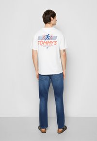 Hvid kortærmet t-shirt med "TOMMY'S Sport & Social Club" trykt i rød og blå på ryggen, parret med blå denim jeans.