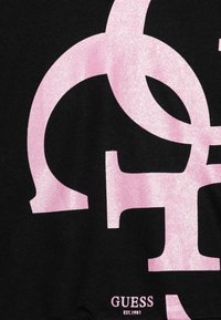 Czarna tkanina z dużymi, błyszczącymi różowymi wzorami logo. Zawiera napis "GUESS EST.1981" w małej czcionce w prawym dolnym rogu.
