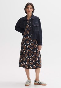 Denim jacket over a patterned, knee-length dress with brown and cream circular motifs; paired with light sneakers. Smooth textures, casual design.
