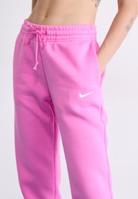Rosa joggebukser med elastisk midjebånd, snøring og sidelommer. Iøynefallende hvit Nike-logo på venstre bein. Myk stofftekstur.