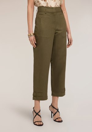 Mujer con pantalones cortos verde oliva con bolsillos delanteros de parche, dobladillo vuelto, sandalias negras de tacón alto con tiras y pulseras doradas en la muñeca.