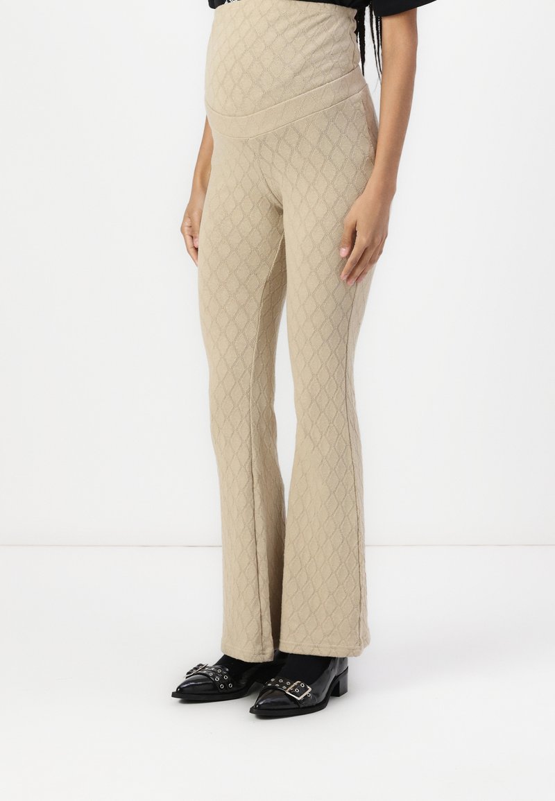Beige flagrende bukser med et struktureret diamantmønster, der har en bred talje og blødt materiale, parret med sorte spidse sko.