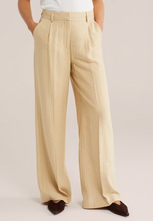 Personne portant un pantalon beige à larges bandes fines avec les mains dans les poches et des chaussures pointues foncées sur un fond neutre.