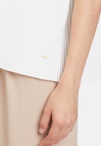 Top bianco plissettato con una targhetta del logo dorato, abbinato a un fondo beige testurizzato, che mette in mostra un tessuto liscio e linee pulite.