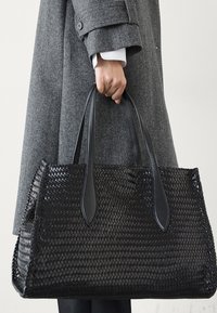 Sac fourre-tout en toile noire avec des poignées en cuir lisse. Présente une forme rectangulaire, une surface texturée et des coutures latérales visibles.
