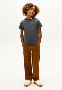 Barn med krøllet hår iført en mørkegrå Tommy Hilfiger t-shirt, brune bukser og hvide sneakers, stående med hænderne i lommerne.