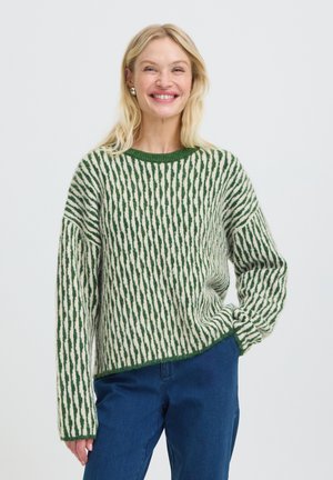 Femme souriante aux cheveux blonds portant un pull vert et blanc à motifs et un jean bleu, debout, main dans la poche, sur un fond uni.