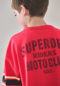 Rød bomuldst-shirt med sorte og cremefarvede striber på ærmerne og fed trykt tekst på ryggen, der lyder "SUPERDRY RIDERS MOTO CLUB 500cc."