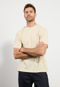 Camiseta de manga corta color beige claro con una sutil textura a rayas, escote redondo y corte clásico, combinada con jeans oscuros.