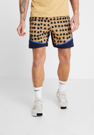 Mann trägt beige Shorts mit großen schwarzen Punkten und blauem Rand, weiße Nike-Socken und weiße Sportschuhe, steht vor einfachem Hintergrund.