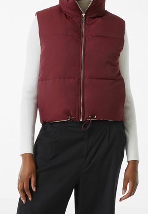 Veste sans manches en doudoune bordeaux avec col montant, fermeture éclair avant et cordon de serrage à l'ourlet. Portée par-dessus un haut blanc à manches longues et un pantalon noir.